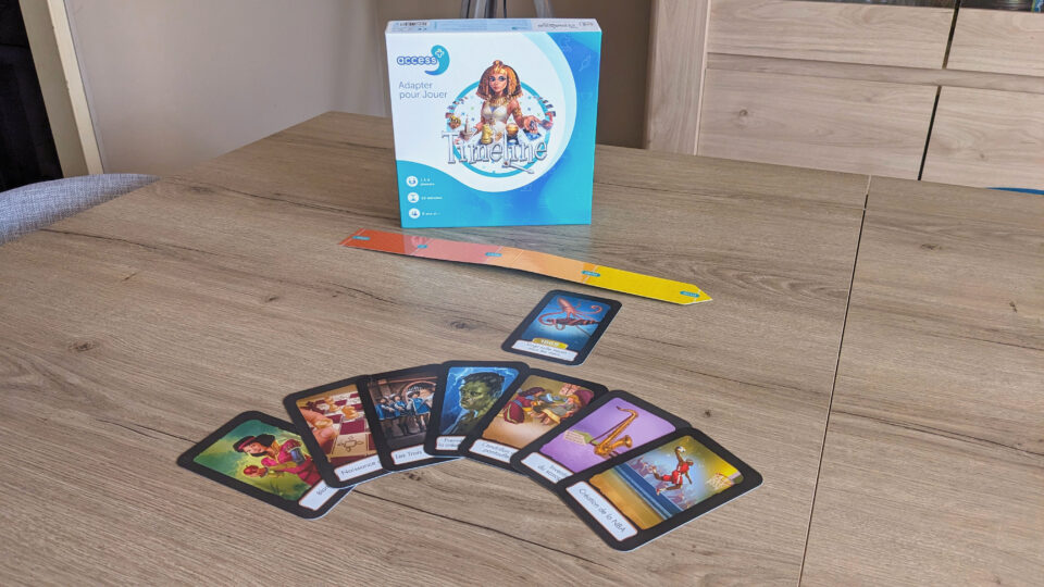 Photo des éléments du jeu Timeline Access+, avec la boîte en fond et la frise chronologique allant de de l'orange au jaune et des dates -8000 à 2010. Quelques cartes du jeu sont également présentes au premier-plan. 